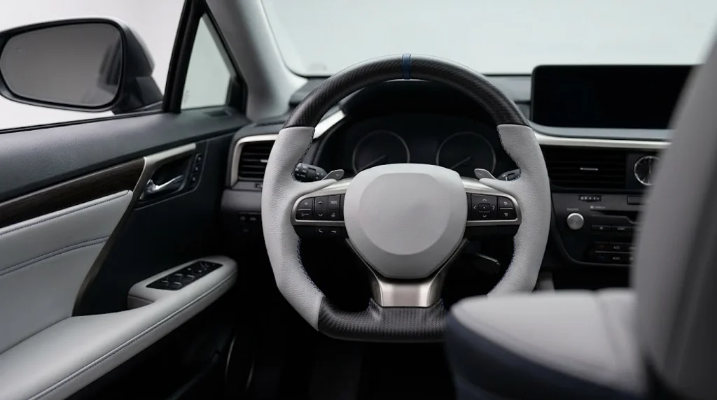 Interior view of an AKIUMI custom steering wheel for Lexus RX, with matte carbon fiber trim, light grey semi-aniline leather grips and deep blue contrast stitching